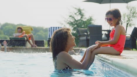 Mother-And-Daughter-Have-Fun-In-Outdoor-Swimming-Pool-On-Summer-Vacation-With-Girl-Sitting-On-Edge