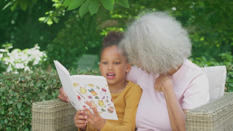 Großmutter-Sitzt-Draußen-Im-Garten-Mit-Enkelin-Zu-Hause-Und-Liest-Gemeinsam-Ein-Buch