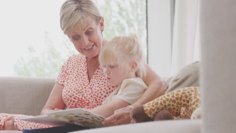Grandmother-Sitting-On-Sofa-With-Granddaughter-At-Home-Reading-Book-Together