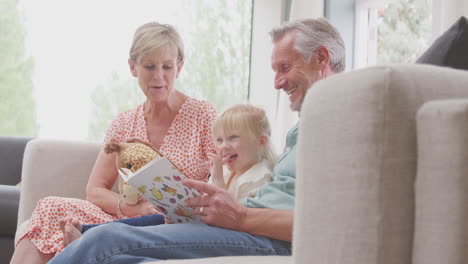 Grandparents-Sitting-On-Sofa-With-Granddaughter-At-Home-Reading-Book-Together