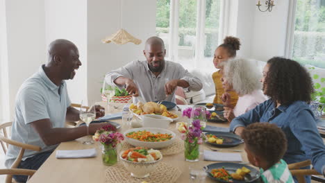Padre-Tallando-Mientras-Una-Familia-Multigeneracional-Se-Sienta-Alrededor-De-La-Mesa-En-Casa-Y-Disfruta-Comiendo