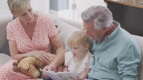 Großeltern-Sitzen-Mit-Enkelin-Zu-Hause-Auf-Dem-Sofa-Und-Lesen-Gemeinsam-Ein-Buch