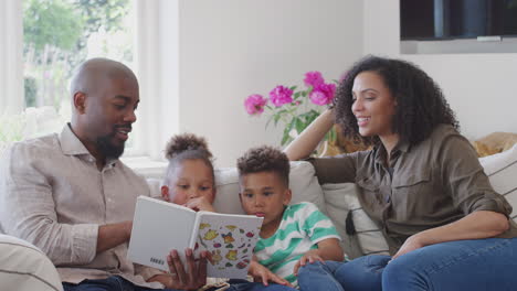 Eltern-Sitzen-Mit-Kindern-Zu-Hause-Auf-Dem-Sofa-Und-Lesen-Gemeinsam-Ein-Buch