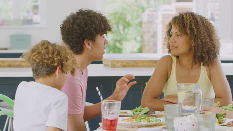 Multi-Racial-Family-Sitting-Around-Table-In-Kitchen-At-Home-Eating-Meal-Together