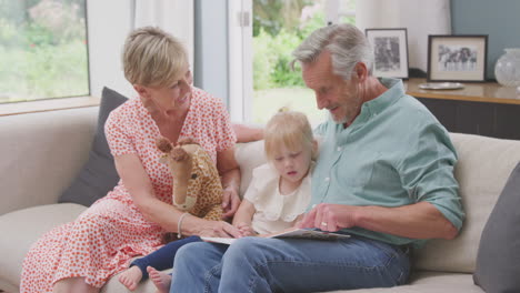 Großeltern-Sitzen-Mit-Enkelin-Zu-Hause-Auf-Dem-Sofa-Und-Lesen-Gemeinsam-Ein-Buch