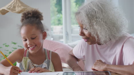 Großmutter-Hilft-Enkelin-Beim-Heimunterricht,-Die-Mit-Einem-Digitalen-Tablet-Am-Tisch-Sitzt