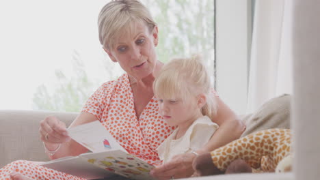 Grandmother-Sitting-On-Sofa-With-Granddaughter-At-Home-Reading-Book-Together