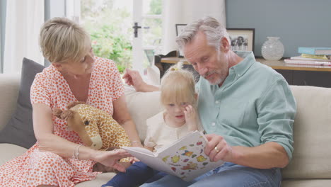Grandparents-Sitting-On-Sofa-With-Granddaughter-At-Home-Reading-Book-Together