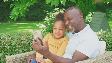 Grandfather-With-Granddaughter-Taking-Selfie-On-Mobile-Phone-In-Garden-At-Home