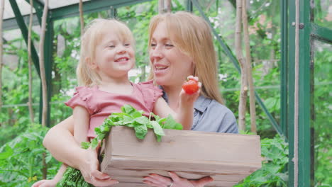 Mother-And-Daughter-Holding-Box-Of-Home-Grown-Vegetables-In-Greenhouse