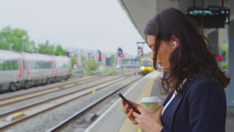 Businesswoman-Waiting-On-Train-Platform-With-Wireless-Earbuds-Answers-Call-On-Mobile-Phone