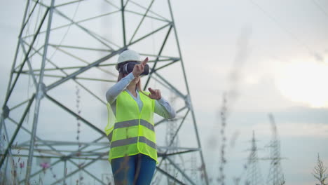 Middle-plan-female-energy-engineer-in-virtual-reality-glasses-and-white-helmet-on-the-background-of-high-voltage-power-line-towers