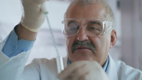 Rack-focused-shot-of-male-Caucasian-scientist-in-medical-gown-and-goggles-pouring-red-liquid-into-test-tube-with-pipette-and-looking-at-result.-High-quality-4k-footage