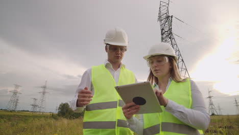 An-electrician-male-and-female-in-the-fields-near-the-power-transmission-line.-He-is-an-electrician-who-manages-the-process-of-erecting-power-lines.-The-mechanic-in-a-helmet-and-Manager-with-tablet