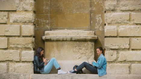Two-Female-University-Or-College-Students-Sitting-Against-Wall-In-City-Working-On-Digital-Tablet