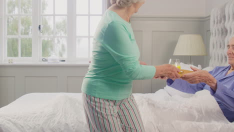 Senior-Couple-At-Home-With-Woman-Bringing-Senior-Man-Breakfast-In-Bed-On-Tray