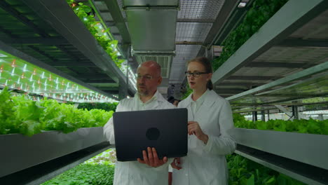 Man-and-a-woman-with-a-laptop-in-white-coats-scientists-go-down-the-corridor-vertical-farm.-Man-and-a-woman-in-white-coats-are-discussing-about-green-plants