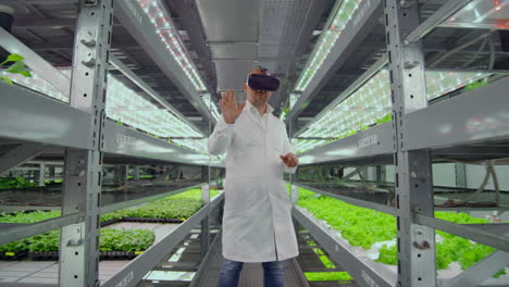 Male-farmer-biologist-in-a-white-robe-standing-in-the-hallway-of-vertical-farming-with-hydroponics-with-glasses-virtualnoy-reality-around-the-green-showcases-with-vegetables.