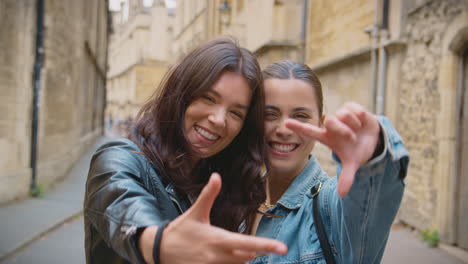 Same-Sex-Female-Couple-Pose-For-Photo-Making-Shape-Of-Frame-With-Hands-As-They-Visit-City-Together