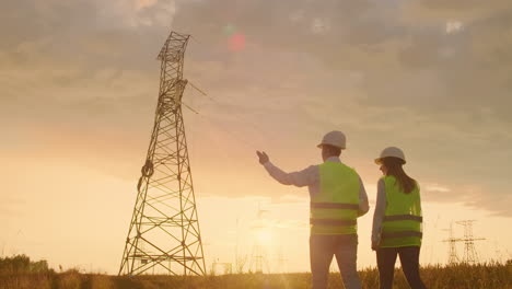 The-view-from-the-back:-Two-engineers-a-man-and-a-woman-in-helmets-with-a-tablet-of-engineer-walk-on-field-with-electricity-towers-and-discuss-the-further-construction-of-towers