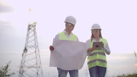 A-group-of-engineers-at-a-high-voltage-power-plant-with-a-tablet-and-drawings-walk-and-discuss-a-plan-for-the-supply-of-electricity-to-the-city.-Transportation-of-renewable-clean-energy.