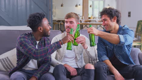 Multi-Cultural-Group-Of-Male-Friends-On-Sofa-At-Home-Drinking-Beer-And-Doing-Cheers-With-Bottles
