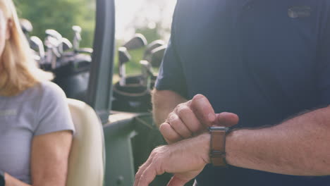 Close-Up-Of-Man-Checking-Smart-Watch-As-Mature-Couple-Sit-In-Buggy-And-Play-Round-Of-Golf-Together