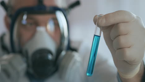 Close-up-in-the-laboratory-a-man-in-a-glass-respirator-screen-holds-a-glass-tube-with-a-blue-liquid-against-the-virus-and-looks-at-it.-High-quality-4k-footage