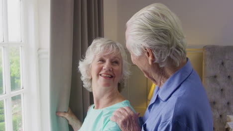 Smiling-Senior-Couple-At-Home-Wearing-Pyjamas-Opening-Bedroom-Curtains-And-Looking-Out-Of-Window