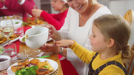 Großmutter-Gießt-Soße-Auf-Das-Essen-Ihrer-Enkelin,-Während-Eine-Familie-Mit-Mehreren-Generationen-Das-Weihnachtsessen-Isst