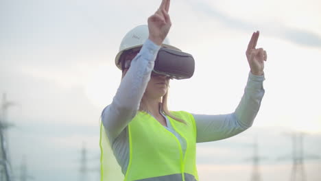 A-woman-electrician-in-virtual-reality-glasses-moves-her-hand-simulating-the-work-with-the-graphical-interface-of-a-power-plant-against-the-background-of-high-voltage-electric-transmission-lines.