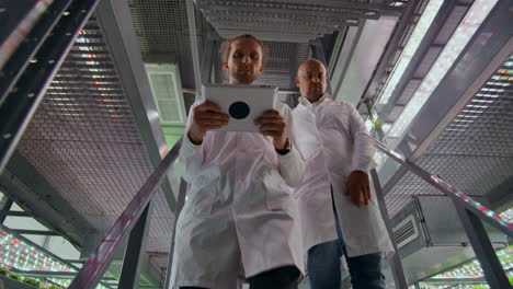 modern-farms-for-growing-organic-products-of-the-future.-two-men-go-down-the-stairs-of-a-vertical-farm-with-a-tablet-computer-in-their-hands-and-2-women-inspect-samples-of-manufactured-products.