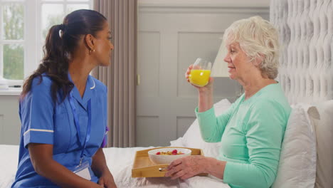 Female-Care-Worker-In-Uniform-Bringing-Senior-Woman-At-Home-Breakfast-In-Bed-On-Tray