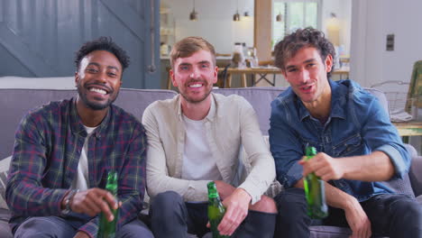 Multi-Cultural-Group-Of-Male-Friends-On-Sofa-At-Home-Drinking-Beer-And-Doing-Cheers-To-Camera