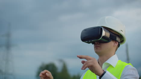 A-male-electrician-in-virtual-reality-glasses-moves-his-hand-simulating-the-work-with-the-graphical-interface-of-the-power-plant-against-the-background-of-high-voltage-electric-transmission-lines.