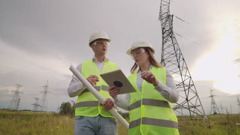 Two-electricians-work-together-standing-in-the-field-near-electricity-transmission-line-in-helmets.-Two-electricians-work-together-standing-in-the-field-near-with-power-transmission-towers.-Eco-friendly-fuel