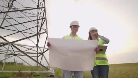 Two-engineers-a-man-and-a-woman-in-helmets-with-a-tablet-of-engineer-walk-on-field-with-electricity-towers-and-discuss-the-further-construction-of-towers
