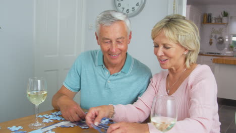 Senior-Retired-Couple-Sitting-At-Table-At-Home-With-Glass-Of-Wine-Doing-Jigsaw-Puzzle