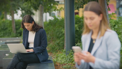 Female-Office-Workers-Outdoors-Working-On-Laptop-And-Using-Mobile-Phone-During-Break-From-The-Office