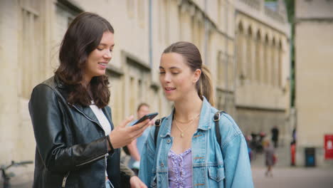 Same-Sex-Female-Couple-Sightseeing-Follow-Map-App-On-Mobile-Phone-As-They-Walk-Around-Oxford-UK
