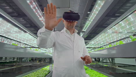 Male-scientist-in-a-white-coat-standing-in-the-hallway-of-vertical-farming-with-hydroponics-with-glasses-virtualnoy-reality-around-the-green-showcases-with-vegetables.