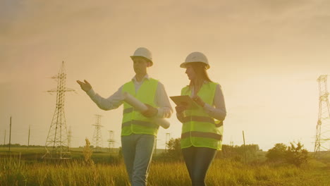 Two-electricians-work-together-standing-in-the-field-near-electricity-transmission-line-in-helmets.-Two-electricians-work-together-standing-in-the-field-near-with-power-transmission-towers.-Eco-friendly-fuel