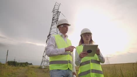 Two-engineers-a-man-and-a-woman-in-helmets-with-a-tablet-of-engineer-walk-on-field-with-electricity-towers-and-discuss-the-further-construction-of-towers