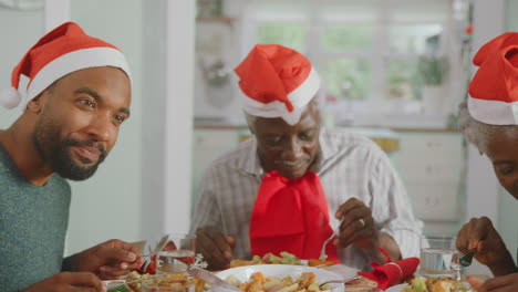 Multi-Generation-Family-In-Santa-Hats-Enjoying-Eating-Christmas-Meal-At-Home-Together