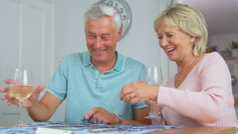 Älteres-Rentnerehepaar-Sitzt-Zu-Hause-Mit-Einem-Glas-Wein-Am-Tisch-Und-Macht-Puzzle
