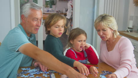 Enkelkinder-Mit-Großeltern-Sitzen-Zu-Hause-Am-Tisch-Und-Machen-Gemeinsam-Puzzles