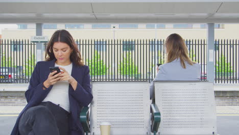Two-Businesswomen-Commuting-To-Work-Waiting-For-Train-On-Station-Platform-Looking-At-Mobile-Phones