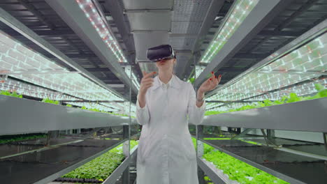 A-woman-in-a-white-robe-standing-in-the-hallway-of-vertical-farming-with-hydroponics-with-glasses-virtualnoy-reality-around-the-green-showcases-with-vegetables.