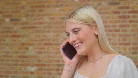 Young-Woman-Standing-By-Brick-Wall-At-Home-Talking-With-Friends-On-Mobile-Phone