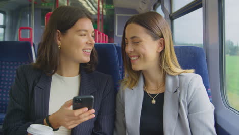Two-Businesswomen-With-Takeaway-Coffees-Commuting-To-Work-On-Train-Looking-At-Mobile-Phone-Together